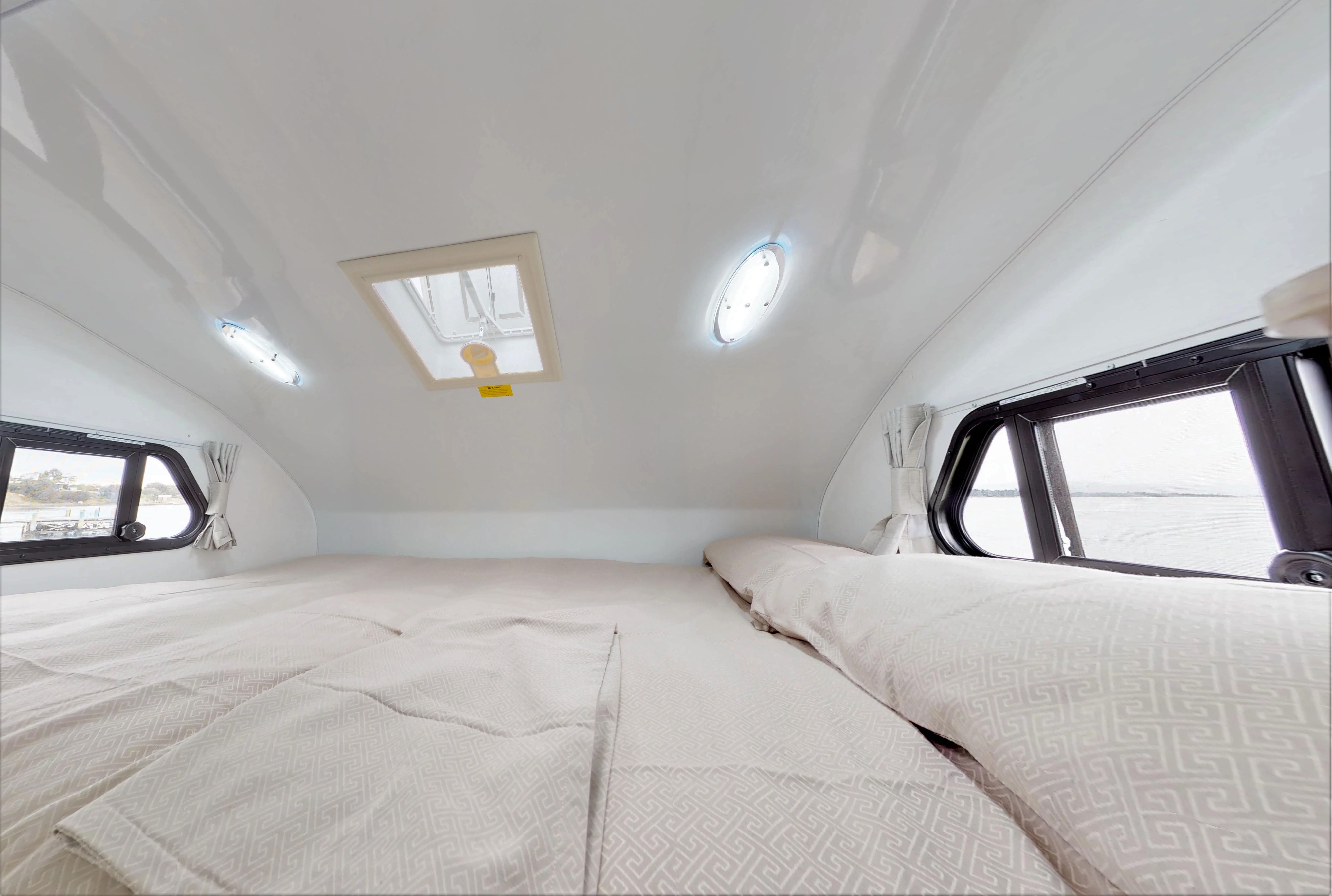 Campervan over-cab bed with beige patterned bedding, curved white ceiling, illuminated lights, and small side windows.