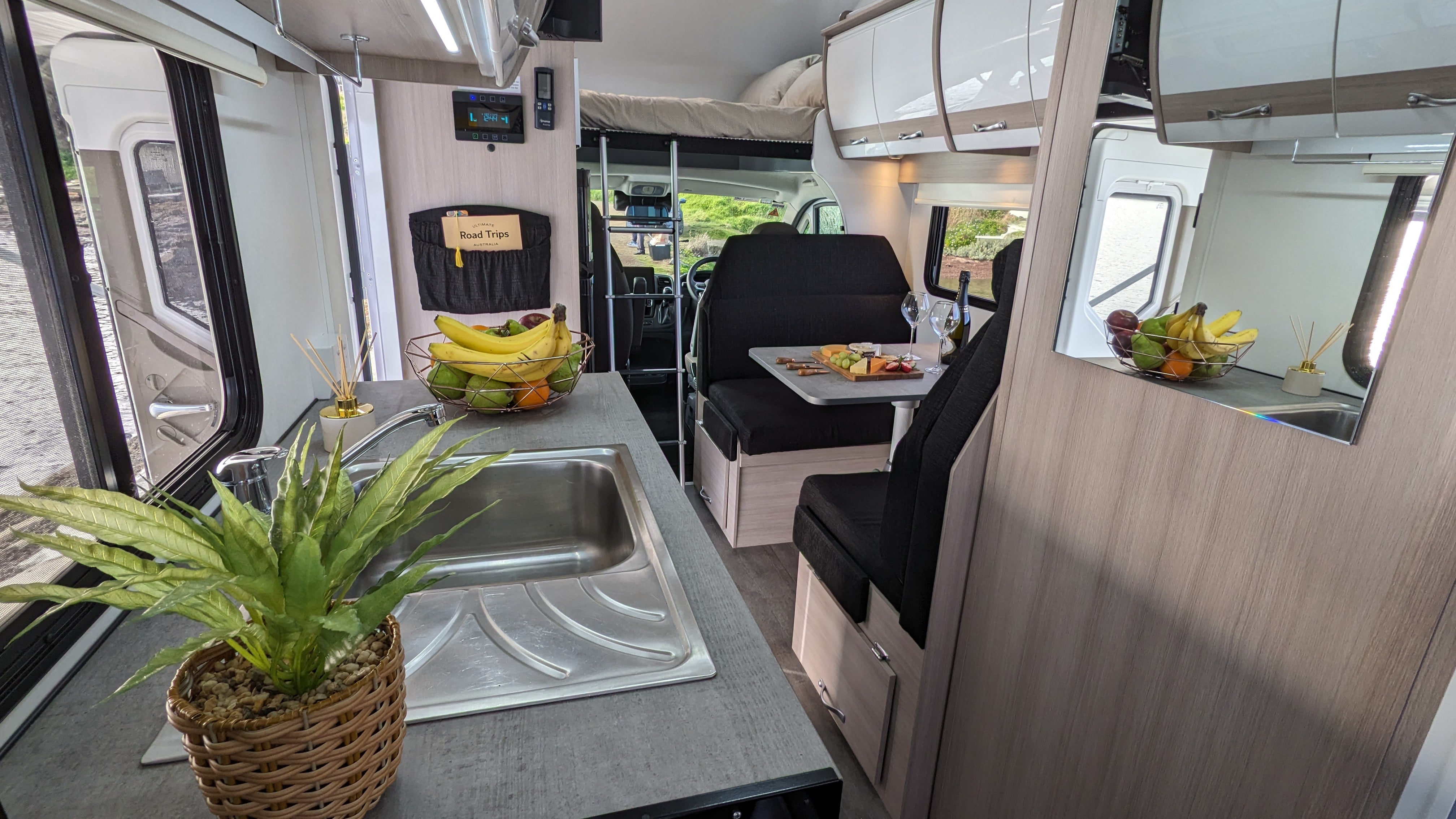 Interior of a modern campervan with a kitchen sink, potted plant, dinette seating area, and an over-cab bed.