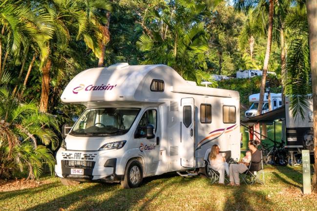 Cruisin' campervan parked at a tropical campsite with two people sitting in folding chairs beside it.