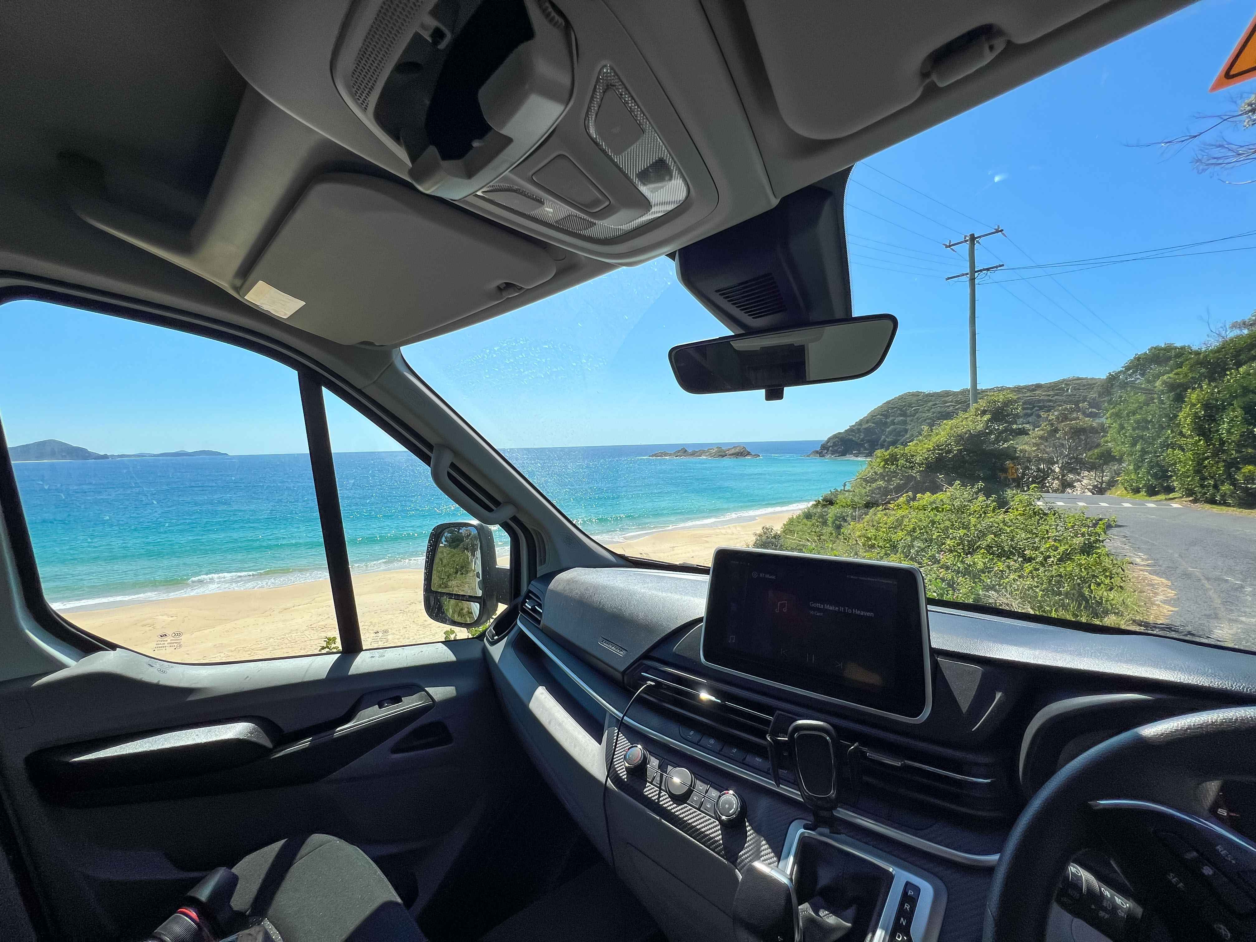 Campervan interior view looking out the windscreen at a sandy beach, turquoise ocean, and a coastal road.