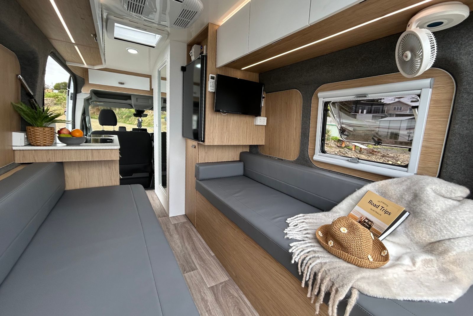 Modern campervan interior featuring grey bench seats, light timber panelling, overhead cabinets, and a window.