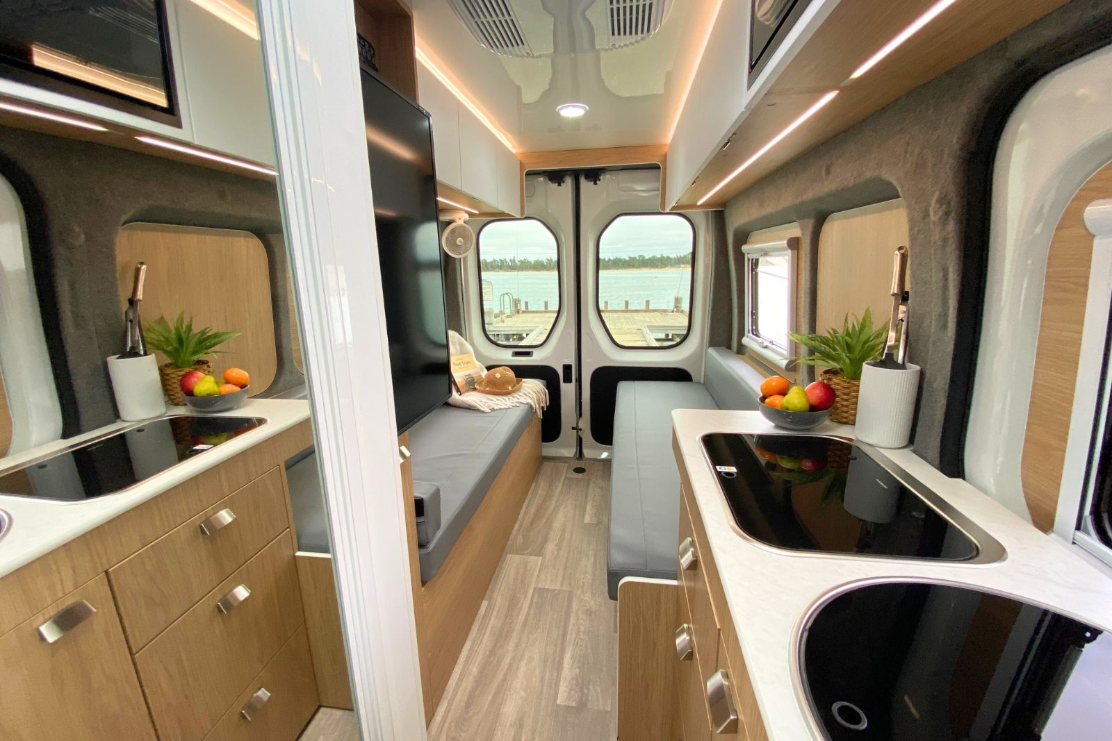 Campervan interior with light timber cabinets, grey bench seats, and water views through the rear windows.