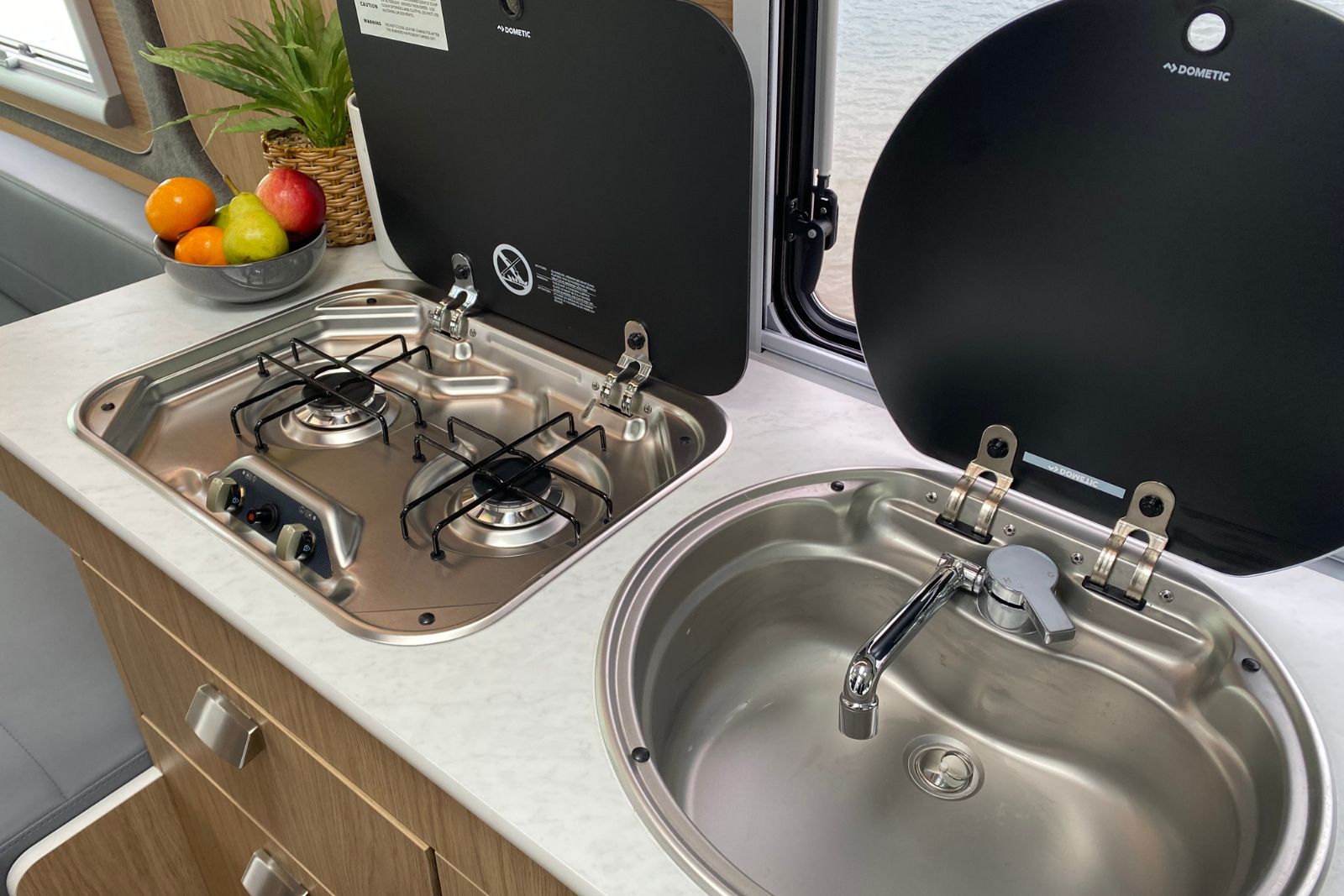 Campervan kitchen benchtop with a stainless steel two-burner gas cooktop and circular sink, both with raised black lids.
