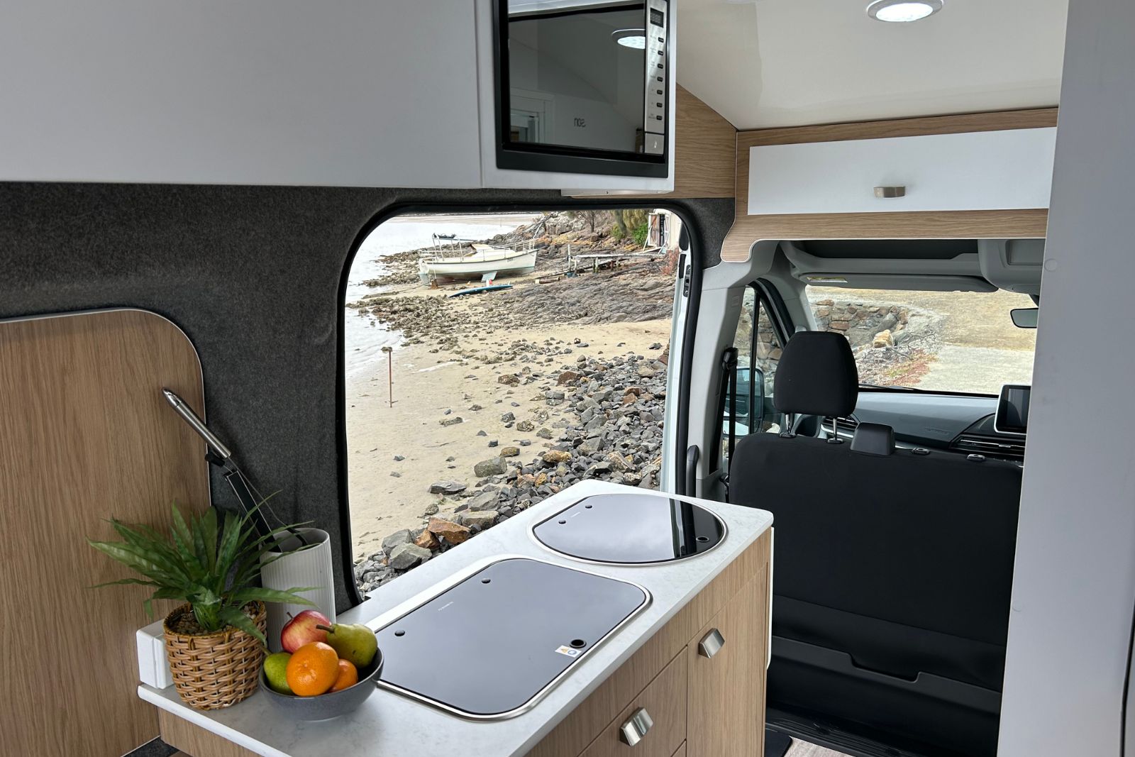 Campervan kitchen counter with sink and stove, looking through the window at a rocky shoreline with a small boat.