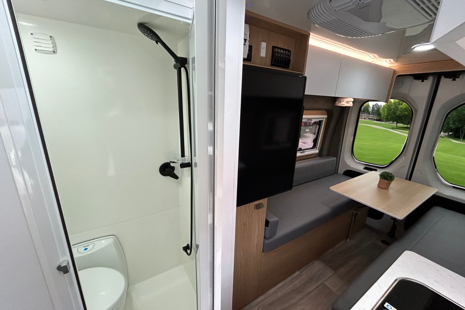 Campervan interior showing a white bathroom with a shower and toilet beside a living area with grey bench seating.