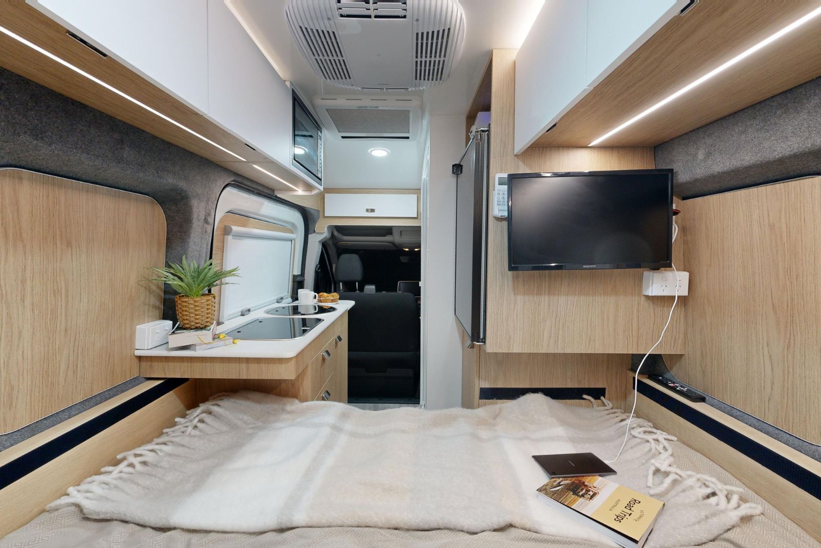 Interior of a modern campervan featuring a bed in the foreground, a compact kitchen, timber panels, and a wall-mounted TV.