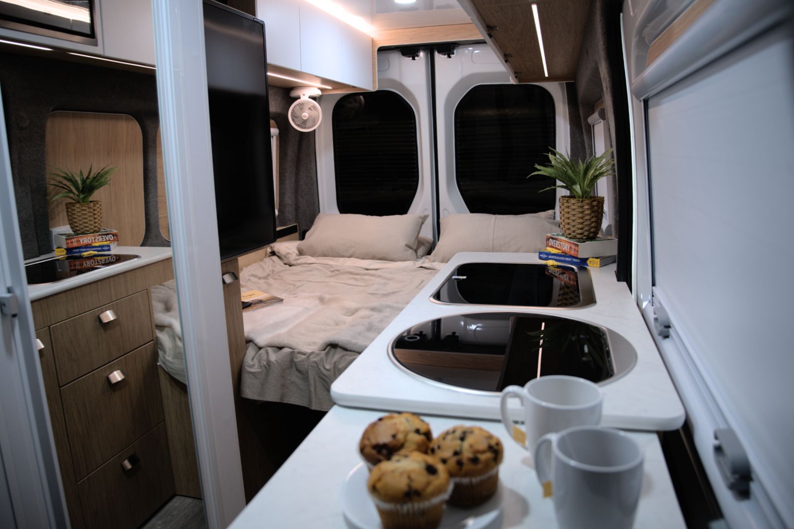 Modern campervan interior with a rear double bed, timber cabinets, and a white kitchen benchtop with muffins and mugs.