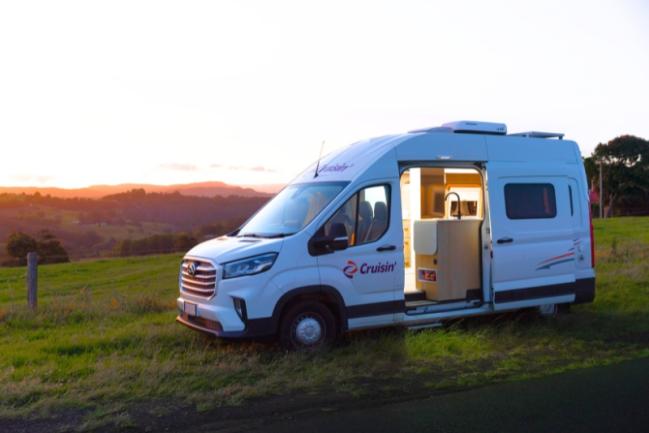 White Cruisin' campervan parked on a grassy hill at sunset with its side door open, revealing a lit kitchen inside.
