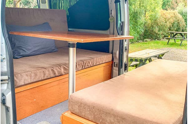 Campervan interior featuring brown cushioned benches, a timber pedestal table, and a view of outdoor picnic tables.