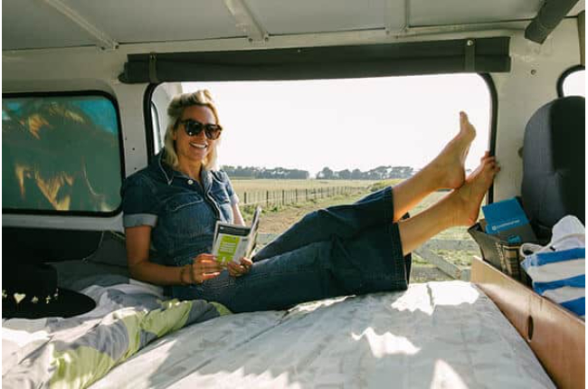 Woman in a denim jumpsuit relaxes inside a campervan, holding a guidebook with her bare feet propped up near the window.