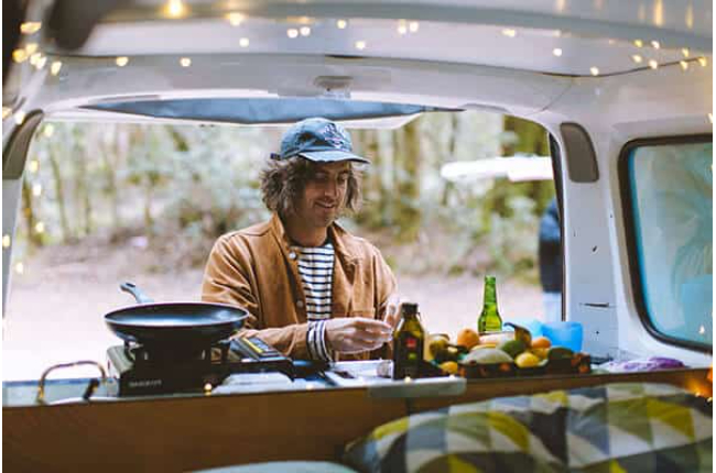 Man preparing food on a portable stove in the back of a campervan decorated with fairy lights.