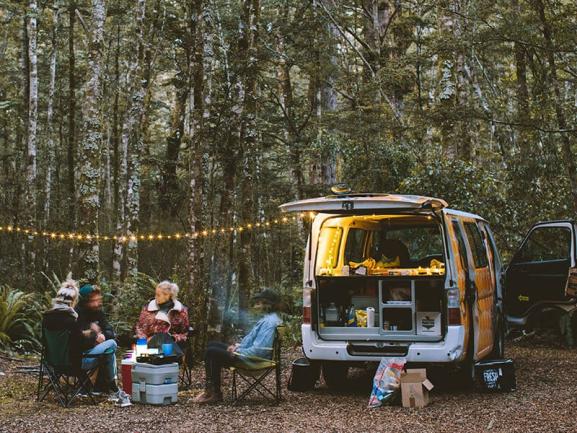 Four people sitting at a campsite in a forest next to an open campervan decorated with string lights.