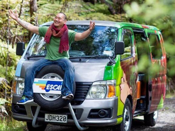 Smiling man with arms outstretched sitting on the front spare tyre of a colourful painted campervan.