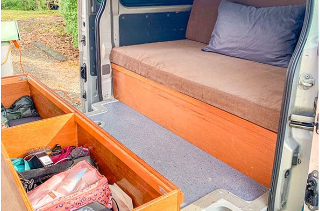 Campervan interior featuring a brown cushioned bed and an open slide-out timber drawer filled with personal items.