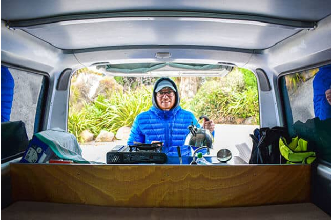 Man in a blue puffer jacket holding a kettle by a stove at the open back of a campervan, viewed from inside.