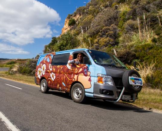 Colourful painted campervan on a road next to a grassy hill, with a woman waving from the driver's window.