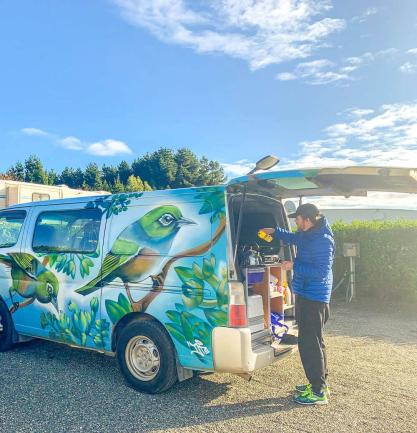 Person using a built-in kitchen at the open back of a campervan painted with a colourful bird mural.