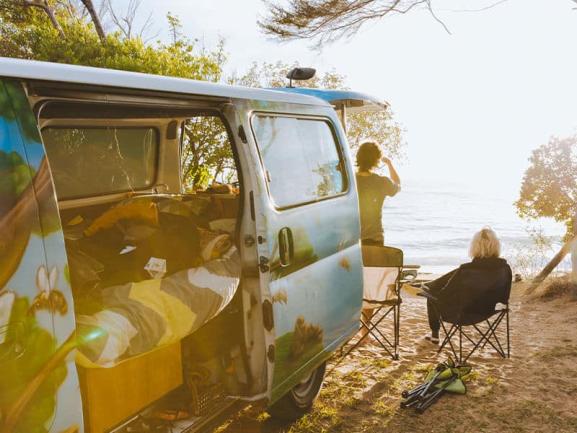 Two people camping beside a painted campervan with its door open, looking out at the ocean.