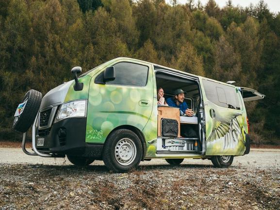 Green campervan with custom graphics parked on gravel, with two people looking out of the open sliding door.