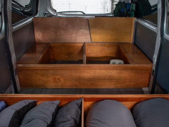 Timber storage benches and open compartments built inside a campervan, with grey cushions in the foreground.