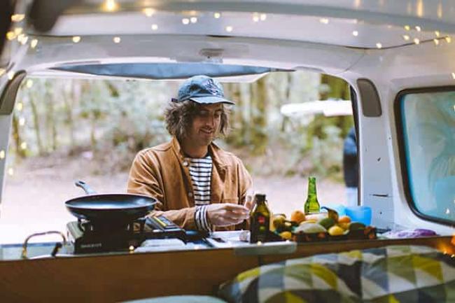 Man prepares food on a portable stove at the back of a campervan, viewed from the interior lined with fairy lights.