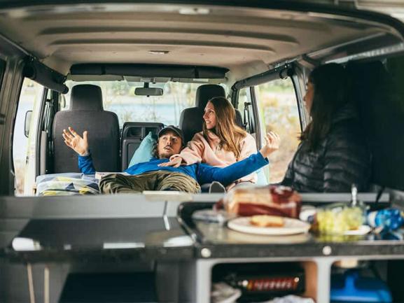 Three friends relaxing and chatting inside a campervan, viewed from behind the kitchen counter.