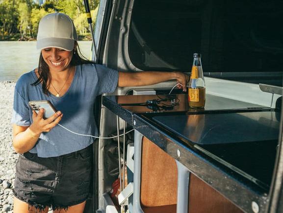 A smiling woman charges her phone from a power outlet inside her campervan kitchen while parked by a river.