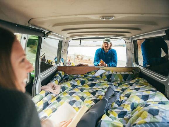 Woman on a bed inside a campervan reading, while a man looks in through the open rear hatch.