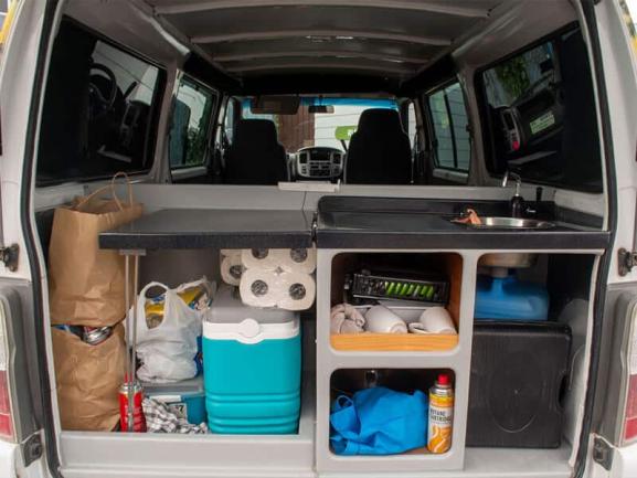 Organised interior of a campervan showing rear storage with a small sink, cooler, shelves, and supplies.
