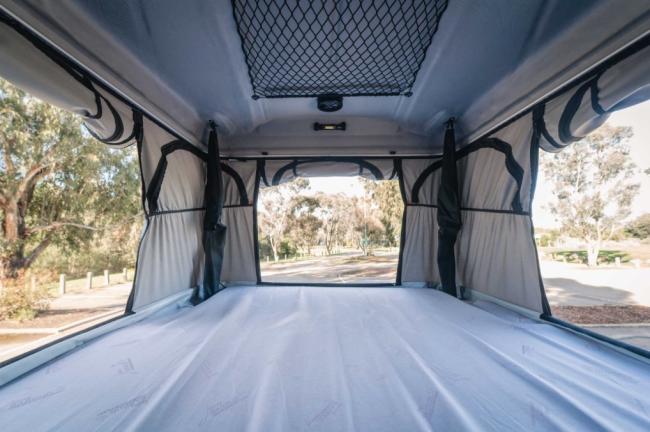 Interior of a campervan pop-top sleeping area with a made bed and open canvas windows looking out at trees.