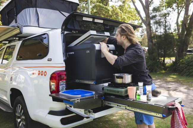 Man using a portable fridge and stove on a slide-out camp kitchen in the back of a white 4WD campervan.