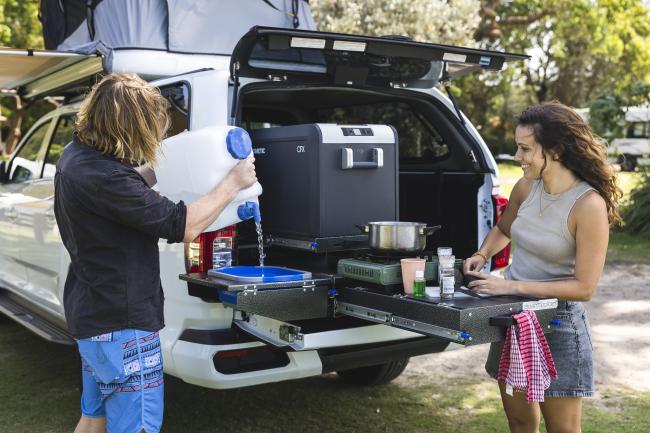 Couple preparing food using a slide-out kitchen system in the back of a white camper vehicle.