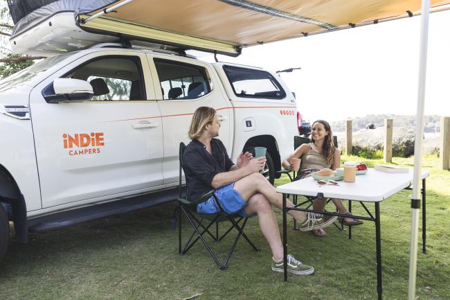 A couple relaxes at a camping table under the awning of a white Indie Campers 4WD utility vehicle.