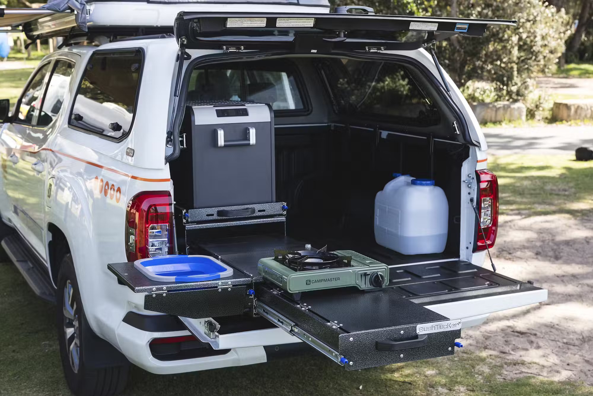 Slide-out camp kitchen in a white ute canopy featuring a portable fridge, gas stove, collapsible sink, and water jug.