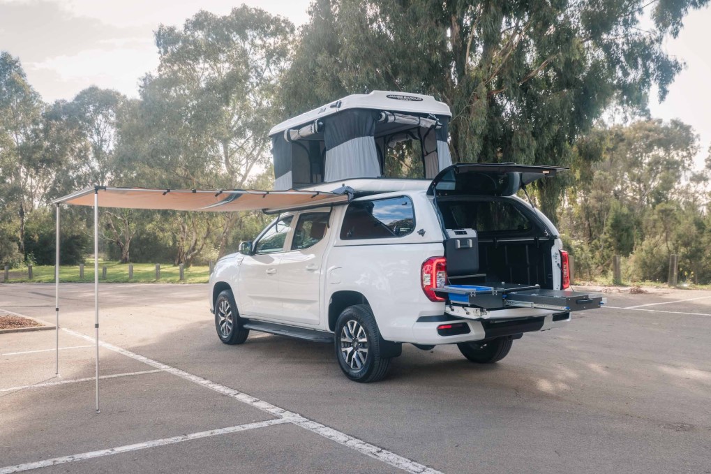 White dual-cab ute with an open rooftop tent, extended side awning, and slide-out kitchen drawer setup.