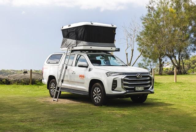 White Indie Campers dual-cab ute with an open rooftop tent and ladder, parked on grass.