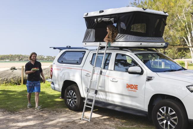 Woman in a rooftop tent on a white Indie Campers 4WD camper while a man walks towards it.