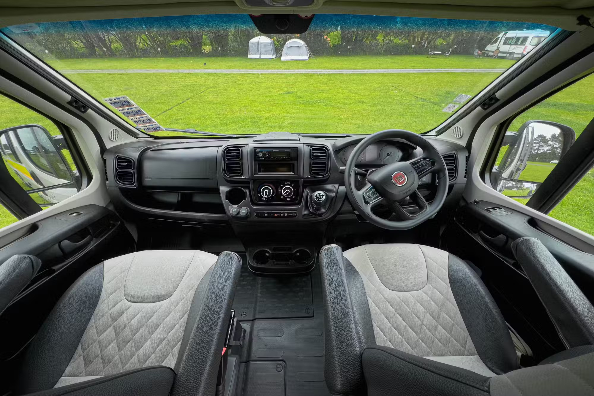 Right-hand drive interior of a Fiat campervan with grey and white quilted seats looking out onto a grassy campsite.
