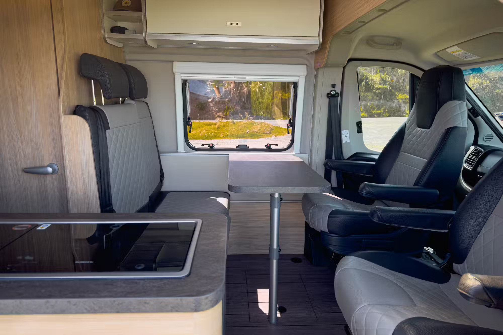 Campervan interior with grey quilted bench and swivel seats around a central dining table, and a window view.
