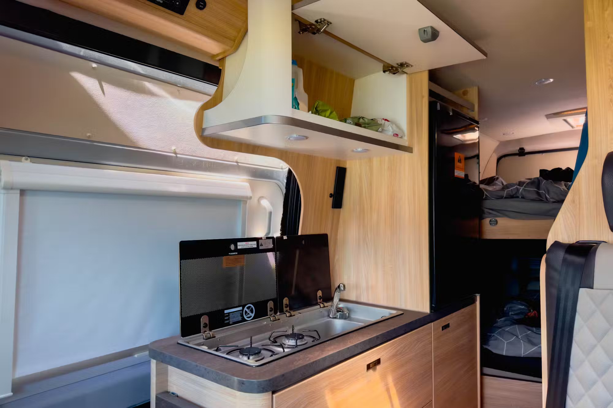 Campervan kitchen with a two-burner stove, sink, timber laminate cabinets, and a fridge next to bunk beds.