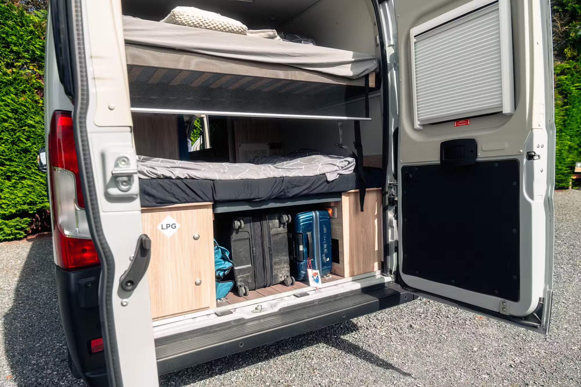 Open campervan rear doors reveal stacked bunk beds and suitcases stored in the under-bed compartment.