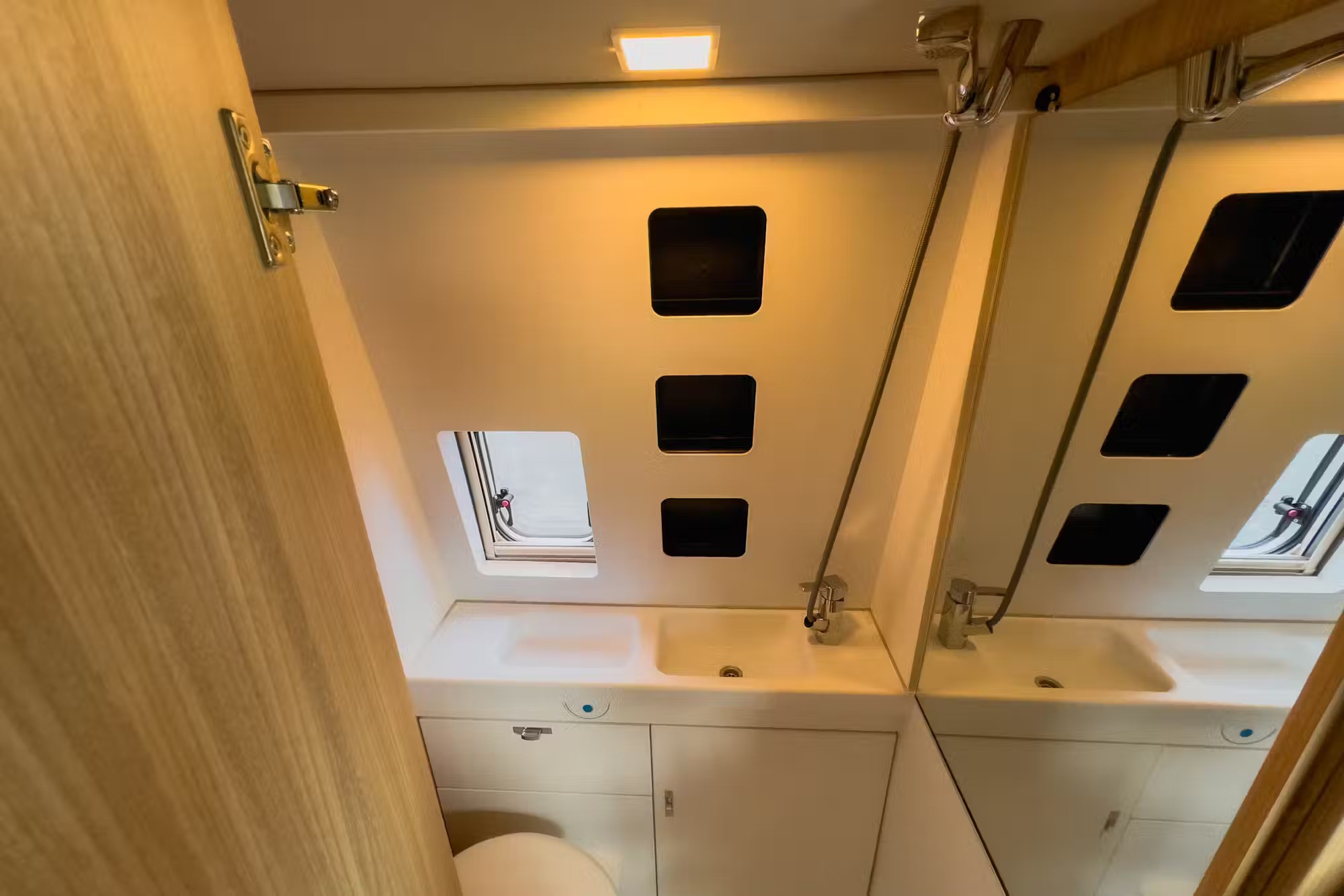 Small campervan bathroom with a white sink, three vertical storage cubbies, a small window, toilet, and large mirror.