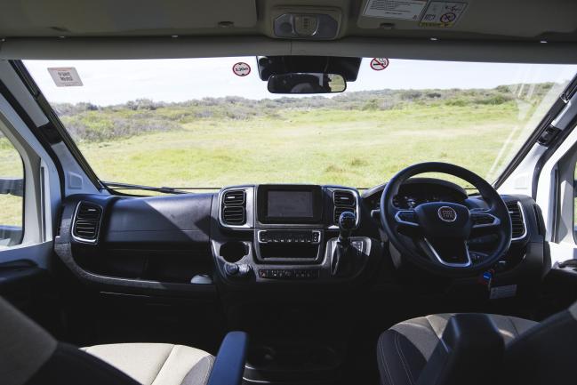 Interior of a right-hand drive campervan cab showing the dashboard and steering wheel looking out onto a grassy field.