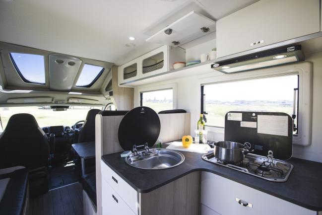Modern campervan interior with a kitchen counter, round sink, gas stove, and front cabin seats in the background.