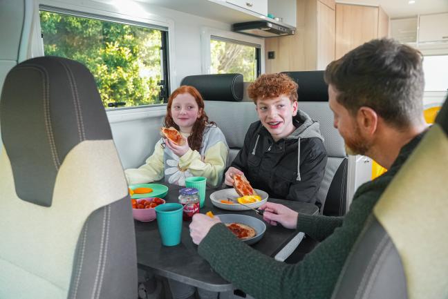 A father and his two children eating toast together at a table inside a campervan.