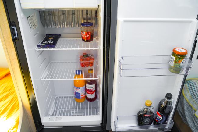 Open compact campervan refrigerator stocked with bottled drinks, jam, chocolate, tomatoes, and condiments.