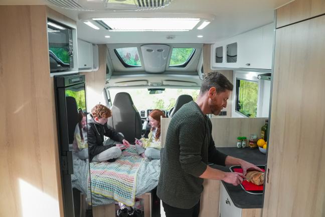 Man slicing bread in a campervan kitchen while two children play cards on a bed in the background.