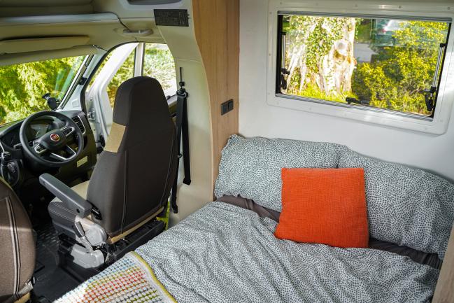 Campervan interior showing a made bed with grey patterned bedding and an orange cushion next to the driver's seat.
