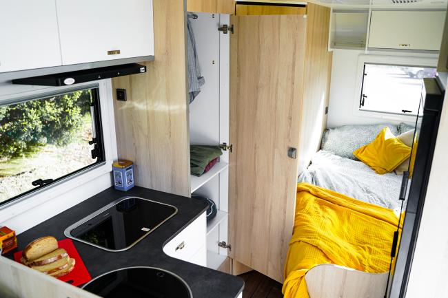 Campervan interior showing a dark kitchen benchtop, light timber-look cabinetry, and a bed with a bright yellow blanket.