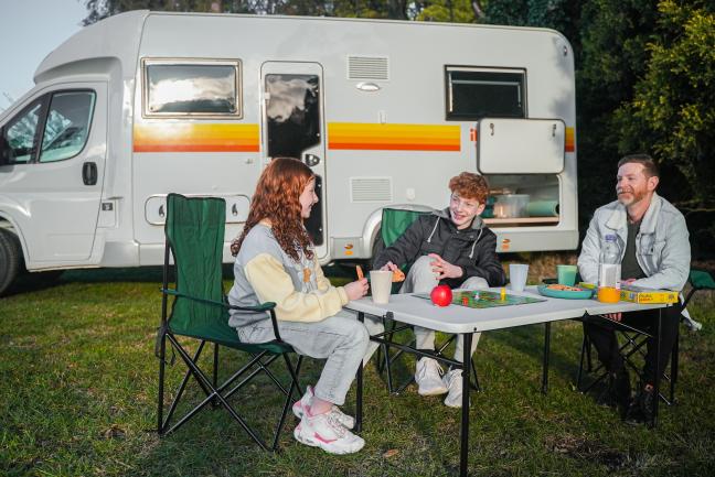 A man and two children sitting at a camp table outside a white campervan, playing a board game on the grass.